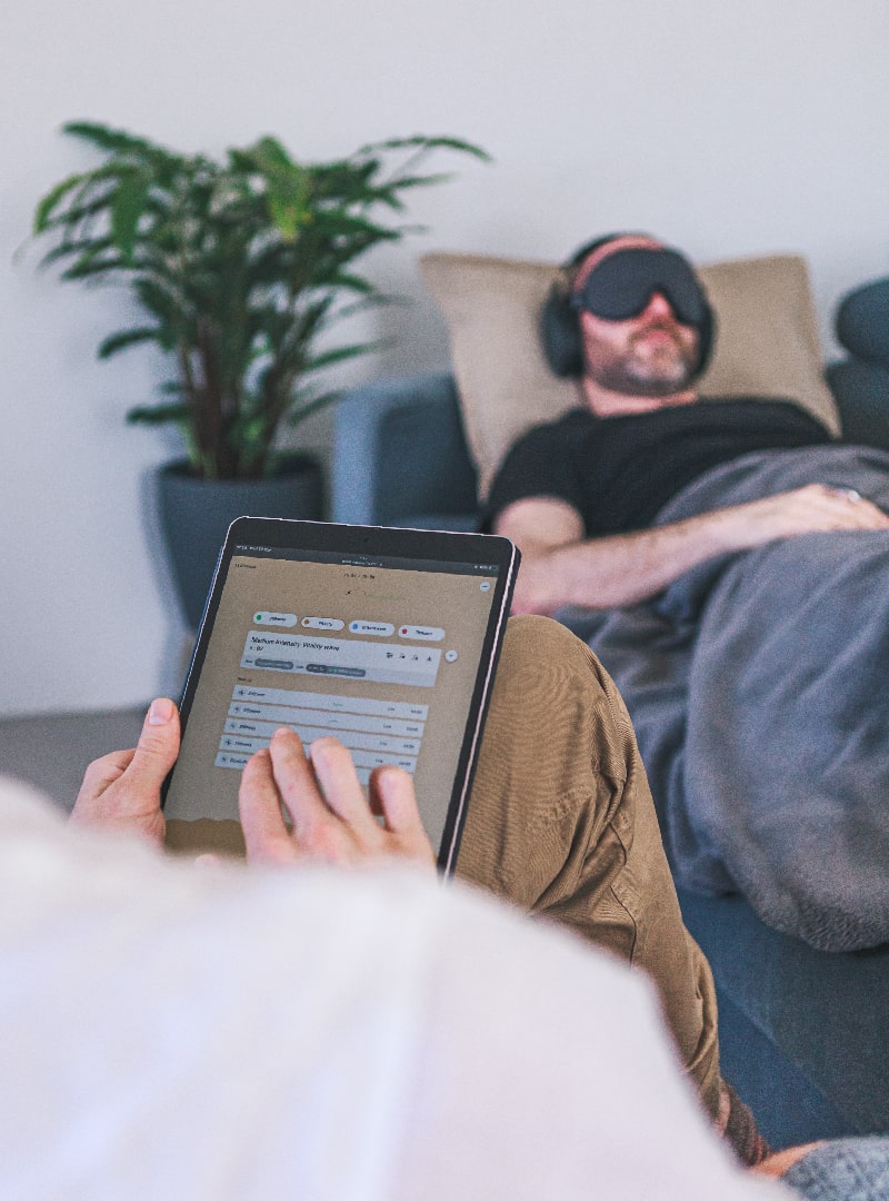 Therapist uses Wavepaths platform on an iPad during Ketamine therapy session. In the background a man is laying down wearing an eye mask and headphones using a blanket.